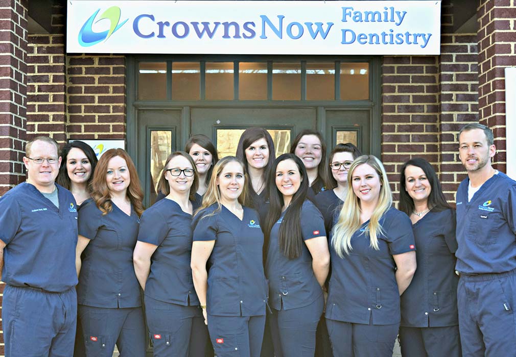 A group of dental professionals posing for a photo in front of a sign that reads  CrownsNow Family Dentistry.