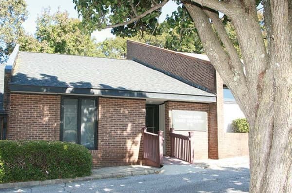 The image shows a single-story brick building with a flat roof, a covered entrance with a glass door, and a parking lot with a tree in the foreground.