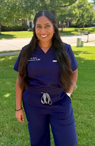 The image shows a woman wearing scrubs standing outdoors with her hands in pockets, smiling at the camera.