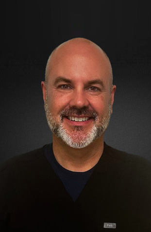 The image shows a man with a beard smiling at the camera  he has short hair and appears to be wearing a dark shirt with a collar.