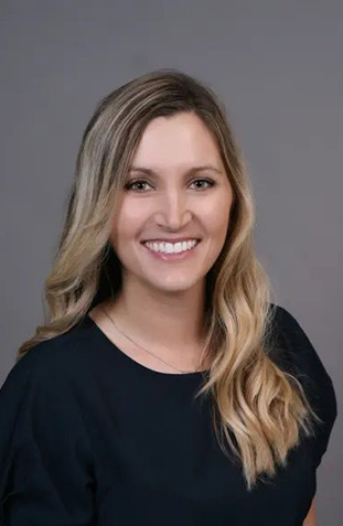 The image shows a woman with long blonde hair wearing a dark top and smiling at the camera, against a neutral background.