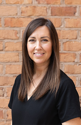 A woman stands confidently against a brick wall, smiling slightly, with short hair and wearing a black top.