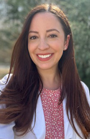 The image shows a woman with long dark hair smiling at the camera, wearing a light-colored blouse and a cardigan, standing outdoors during daylight.