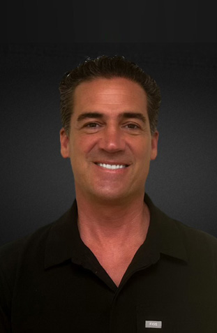 The image shows a smiling man with dark hair, wearing a black shirt, standing against a dark background.