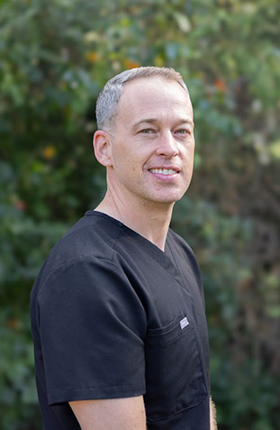 This is a photograph of a man wearing scrubs, standing in front of a brick wall.