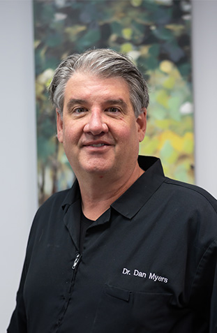 A man in a black apron stands against a white wall, smiling at the camera.