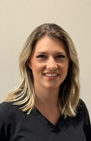 The image is a photograph of a woman with short hair, wearing a dark top and a lanyard. She has a professional appearance, likely in a healthcare or service-related setting, as suggested by her attire and the background that appears to be an indoor office environment.