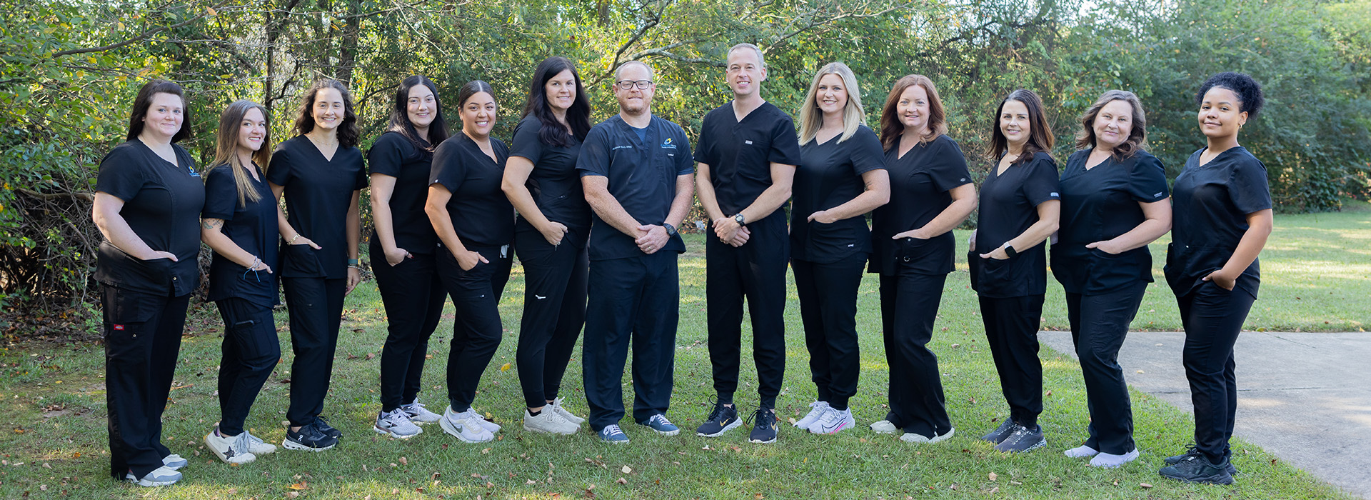The image shows a group of individuals, likely professionals, posing together in front of a building with a sign that reads  DENTAL CLINIC . They appear to be dressed in scrubs and are smiling at the camera.