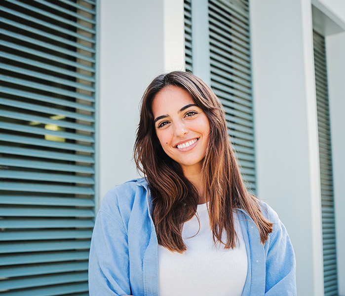 The image is a portrait of a young woman with medium-length hair, smiling at the camera, wearing a light blue top and standing in front of a building with horizontal blinds.