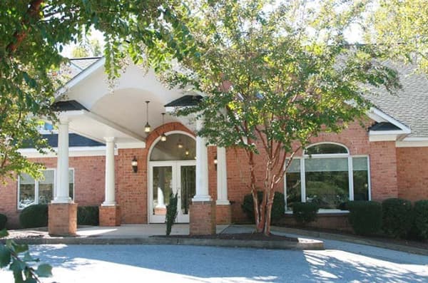 The image shows a large, two-story building with a prominent front entrance. The architecture features a mix of brick and stone facades with white trim around the windows and doorways. There is a covered walkway leading to the entrance, which appears to be designed for accessibility, as indicated by the presence of a ramp. A large arched window with multiple panes spans the front of the building above the entrance. The exterior walls are adorned with landscaping elements, including shrubbery and small trees, contributing to a well-maintained appearance. The sky is visible in the background, suggesting that it is daytime.