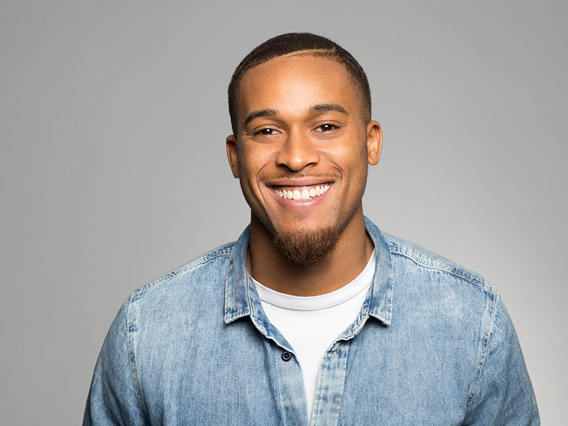 The image features a smiling man with short hair, wearing a denim jacket and a white t-shirt, standing against a plain background.