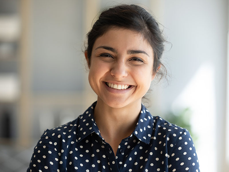 A young woman with a bright smile, wearing a polka dot blouse and a dark jacket, poses confidently against a blurred background.