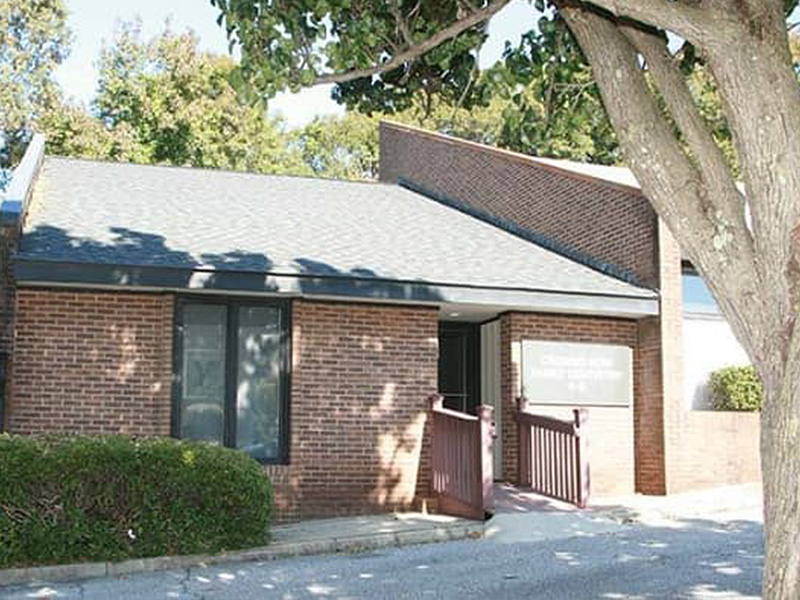 The image shows a single-story brick building with a dark roof, featuring a prominent gabled section and a flat-roofed extension. A small porch is visible on the front of the building, supported by columns. The structure appears to be a church or a community center, situated in a suburban area with trees and grass around it. There are no people present in the image.