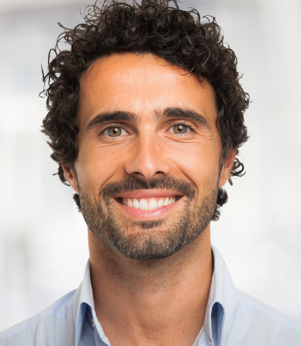 The image shows a man with short hair, smiling at the camera. He has a beard and is wearing a dark blue shirt. His background appears to be an indoor setting with a blurred backdrop that suggests a professional environment.