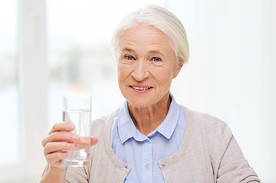 The image shows an elderly woman holding a glass of water, smiling slightly, with a warm and welcoming expression.