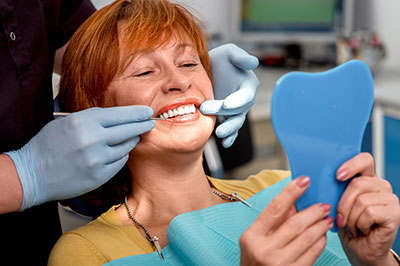 The image shows a woman sitting in a dental chair, receiving dental treatment. She is holding up a blue mouthguard and smiling at the camera while wearing a face mask with clear glasses over her eyes. A dental professional is standing behind her, adjusting her teeth with a tool.