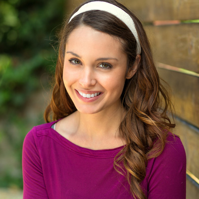 A smiling woman with long brown hair, wearing a purple top and headband, stands against a wooden fence.