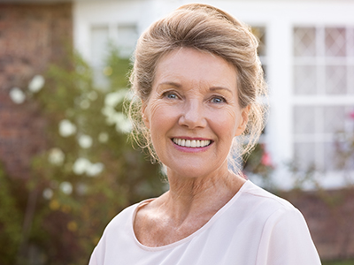 The image shows a woman standing outdoors in front of a house, smiling at the camera. She has short blonde hair and is wearing a light-colored top with a darker collar.