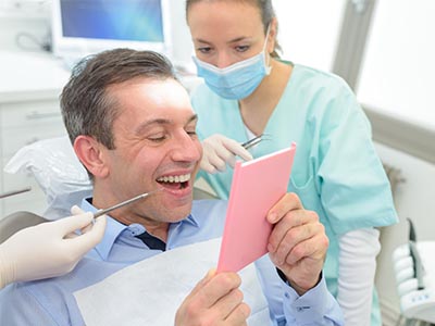 The image shows a man in a dental chair, smiling and holding a pink card, with a female dentist in the background looking at him.