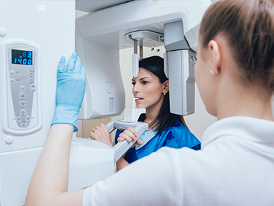 An image featuring a woman in a blue coat standing near a large, modern CT scanner, with another person partially visible on the right side.