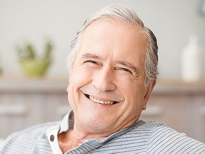 The image shows an elderly man with gray hair, smiling broadly and looking directly at the camera. He is wearing a dark shirt and has his head tilted slightly upwards. The background is blurred but appears to be a domestic setting with a hint of a kitchen counter and cabinets.