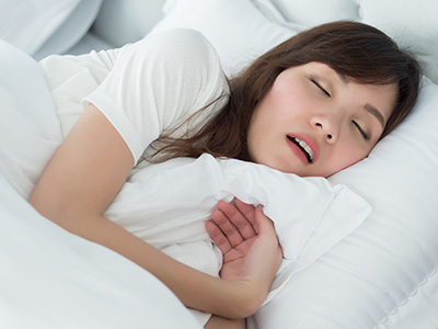 Sleeping woman with closed eyes, wearing a white t-shirt and black pants, on a bed with a white comforter.