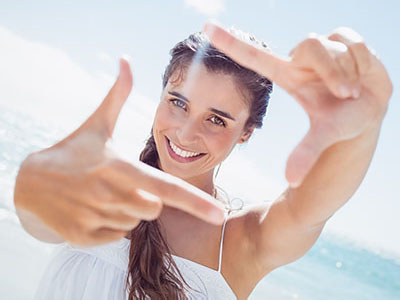 The image shows a woman with her hand in front of her face, making the peace sign, against a beach backdrop.