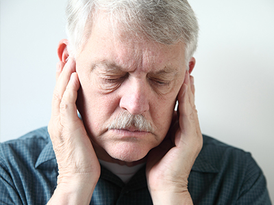 The image shows an older man with a mustache, holding his face with both hands over his ears, as if in pain or discomfort.